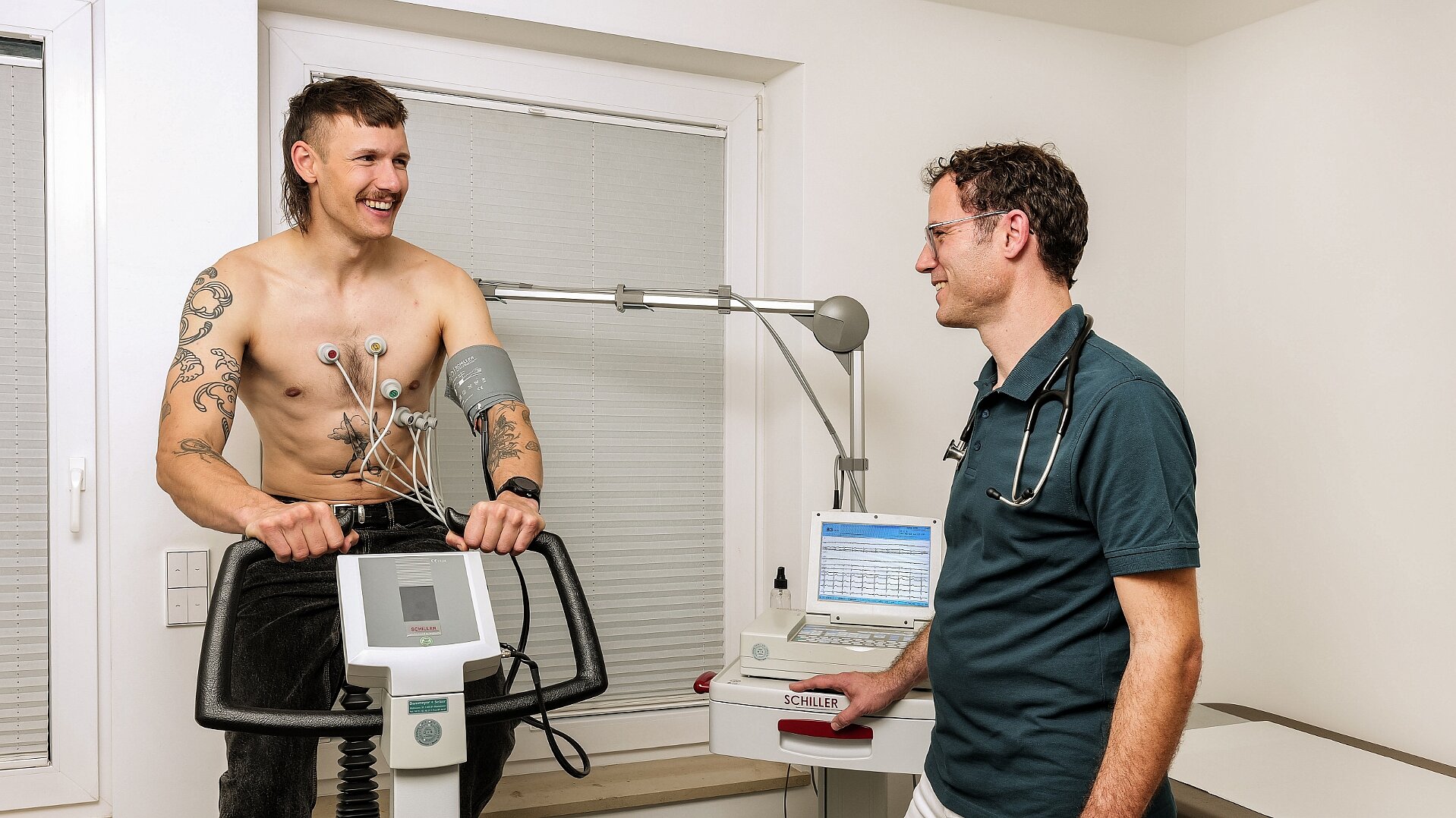 Junger tätowierter Mann während Belastungs-EGK mit begleitendem Arzt rechts von ihm